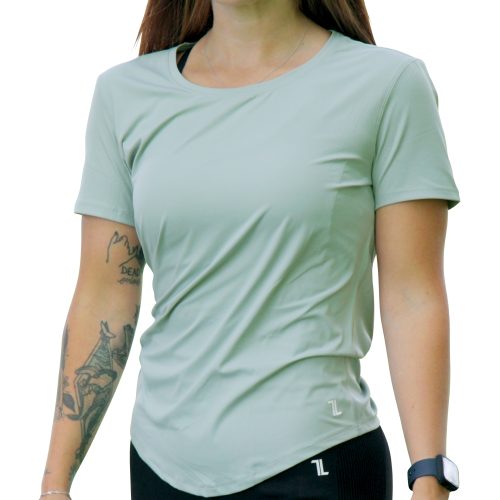 A woman with long brown hair, a left forearm tattoo, and a smartwatch walks outdoors wearing black leggings and the VeloraFlow Tee - Sage Green.